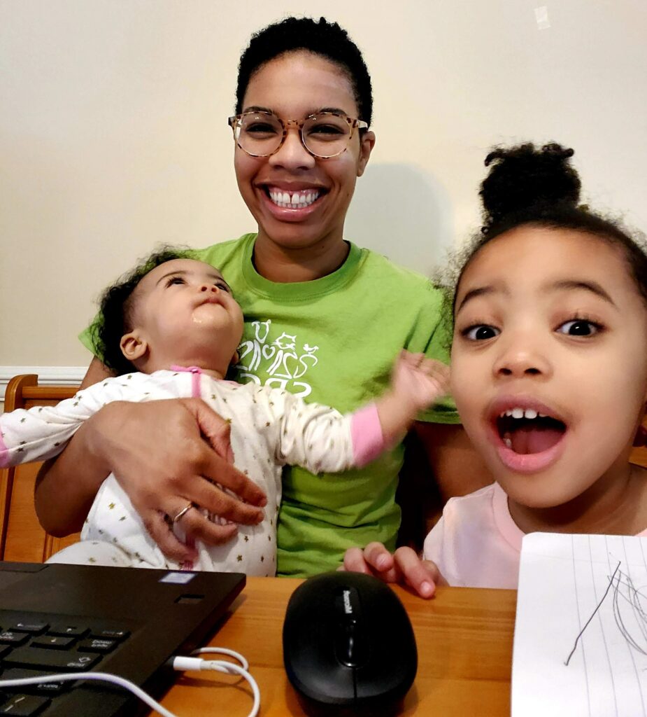 Jessika Jackson smiles with her two daughters pressed against her face. She's wearing a casual T-shirt that is mostly white, her infant daughter in the middle has on a stained white onesie and her daughter at the end has on a white tank top.