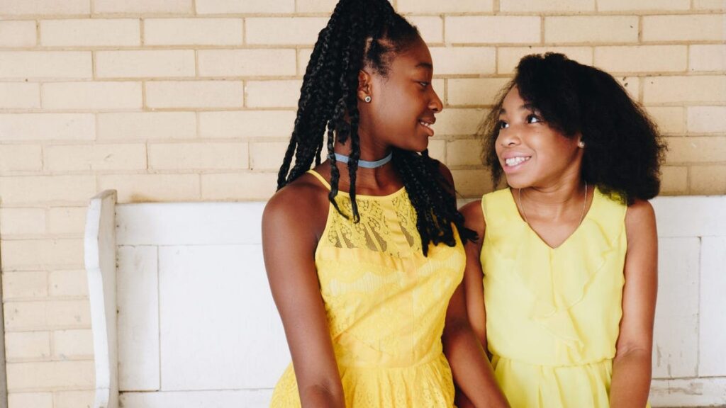 Two Black girls both in sleeveless yellow shirts smile at each other. One wears her hair in braids with beads at the end and the other has a natural, wavy look.
