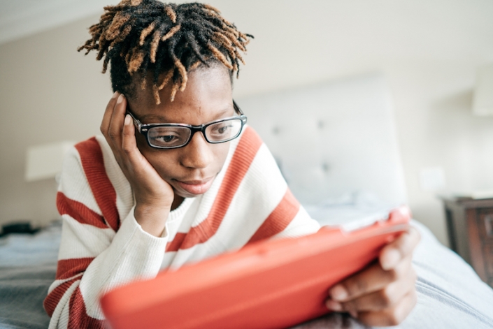 Adolescent Black boy with short dredlocks that have blond tips stares at a tablet with a red cover. He is wearing black-framed glasses and a white sweater with red stripes. 