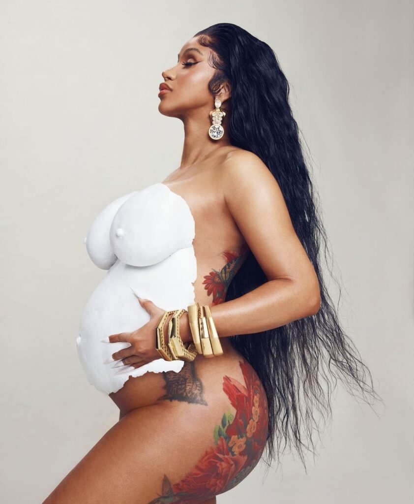 A full-body profile shot of rapper Cardi B posing pregnant with a white belly sculpture. She is wearing long hair and chunky gold bracelets