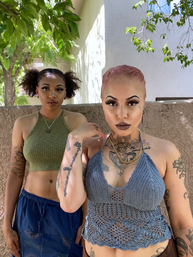 Courtney is pictured standing behind Lex. Courtney has a green halter crop top and two ponytails while Lex has a short slicked back pink hairstyle with a gray crop top. 