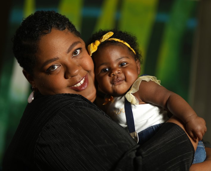 Krista Hayes hold her infant daughter cheek to cheek as they both smile for a portrait against an abstract green background.