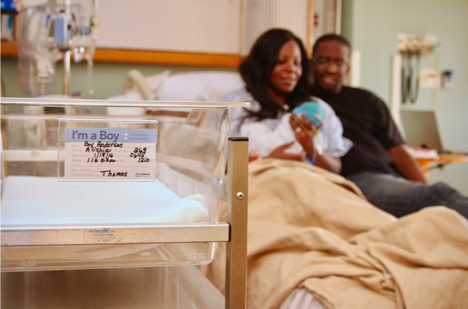 A mother and father hold their child in a hospital bed with a close up of a baby bin.