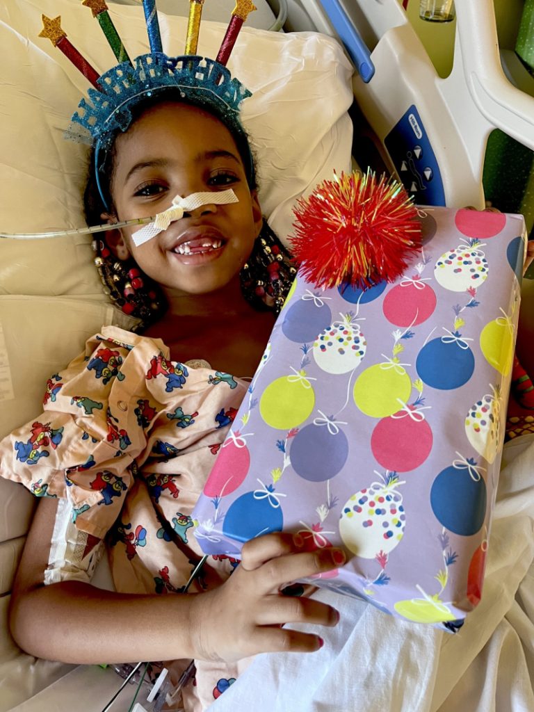 Little girl with NG tube smiles in hospital bed while holding wrapped birthday gift. 