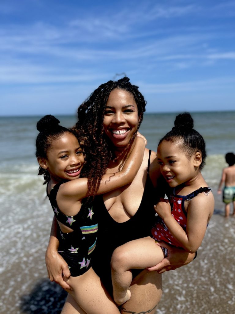 Womanin black bathing suit hold two young children at the beach