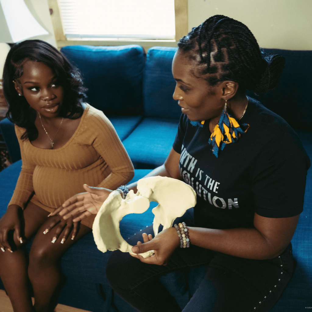 Black doula holds pelvic figure while talking to pregnant Black mom on couch.